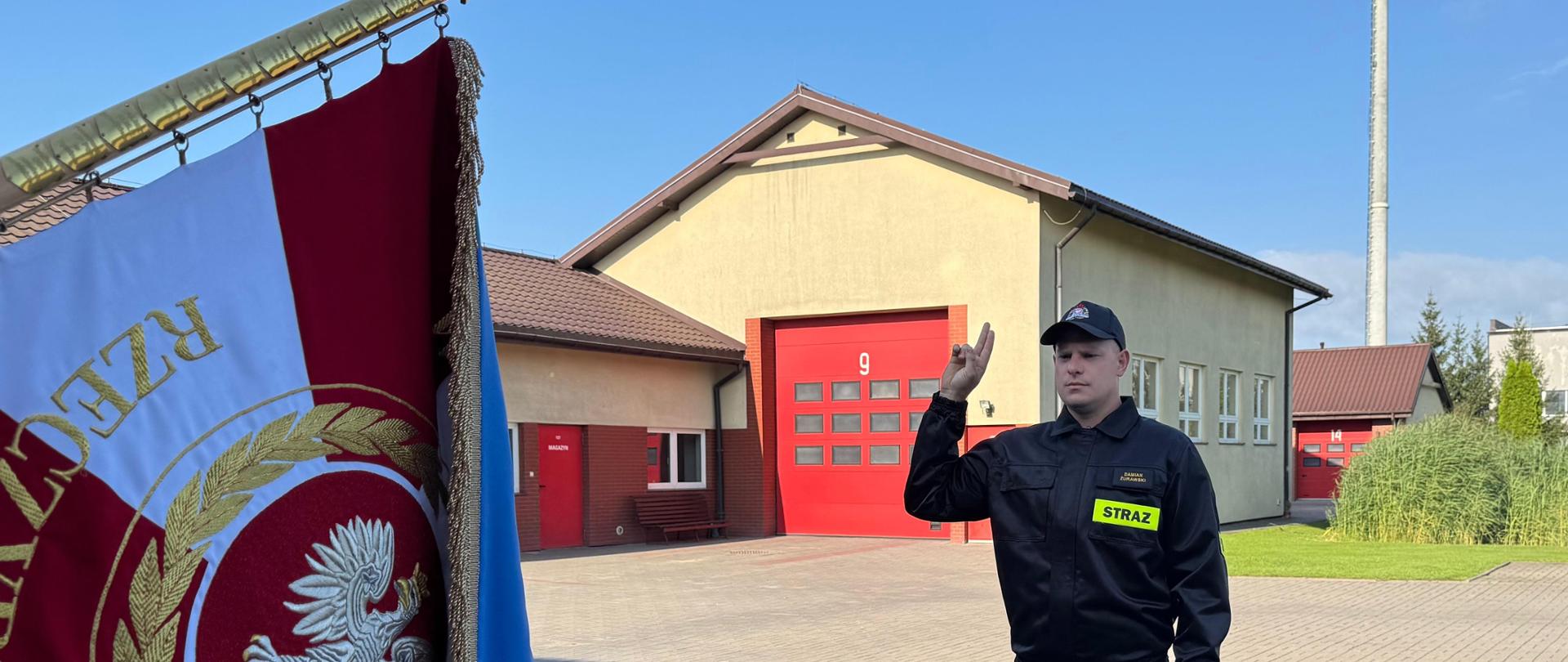 Na pierwszym planie funkcjonariusz Państwowej Straży Pożarnej składa rotę ślubowania podczas uroczystości przyjęcia do służby. W tle widoczne są plac Komendy Powiatowej Państwowej Straży Pożarnej w Rypinie oraz garaże jednostki.