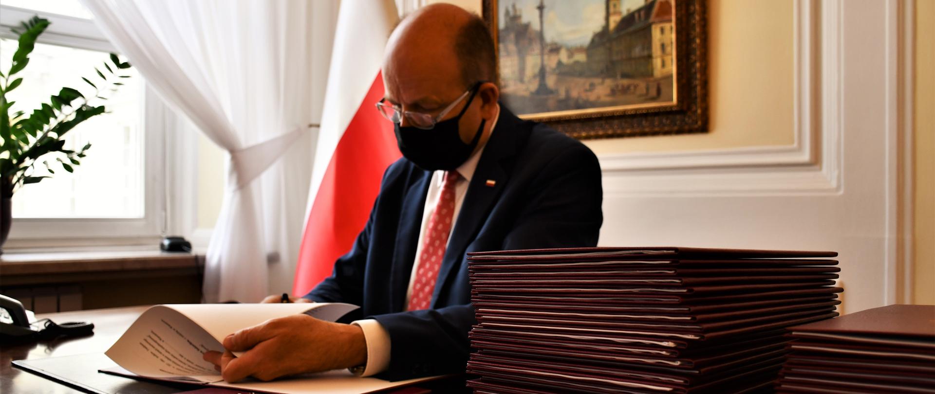 Wojewoda Mazowiecki siedzący za biurkiem. Ubrany w elegancki garnitur podpisuje umowy w ramach Funduszu Solidarnościowego