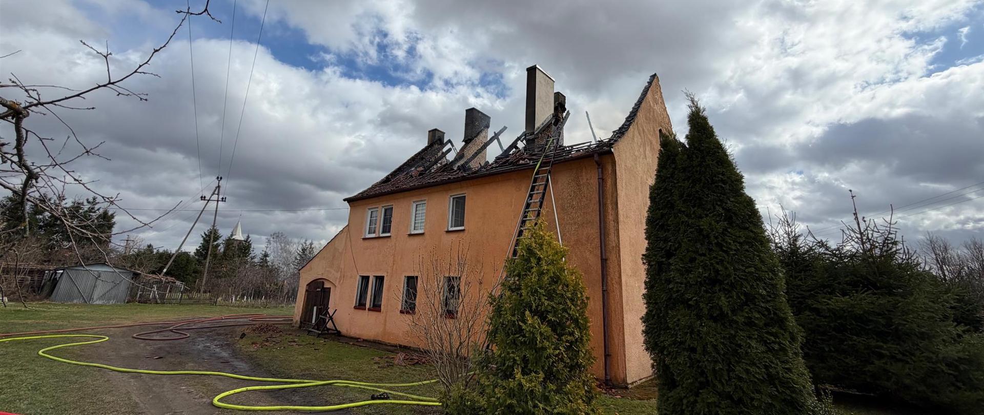 Perspektywa budynku z boku, ukazująca skalę zniszczeń poszycia dachowego. O ścianę domu oparta jest długa drabina strażacka sięgająca kalenicy. Wokół domu na ziemi rozciągnięte są żółte i czerwone węże strażackie.