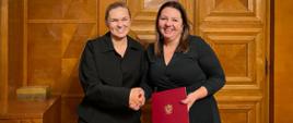 Na zdjęciu widoczna minister Barbara Nowacka oraz Izabela Ziętka. Minister wręcza teczkę z powołaniem i podaje rękę. Kobiety ubrane na czarno stoją na tle drewnianych drzwi. 