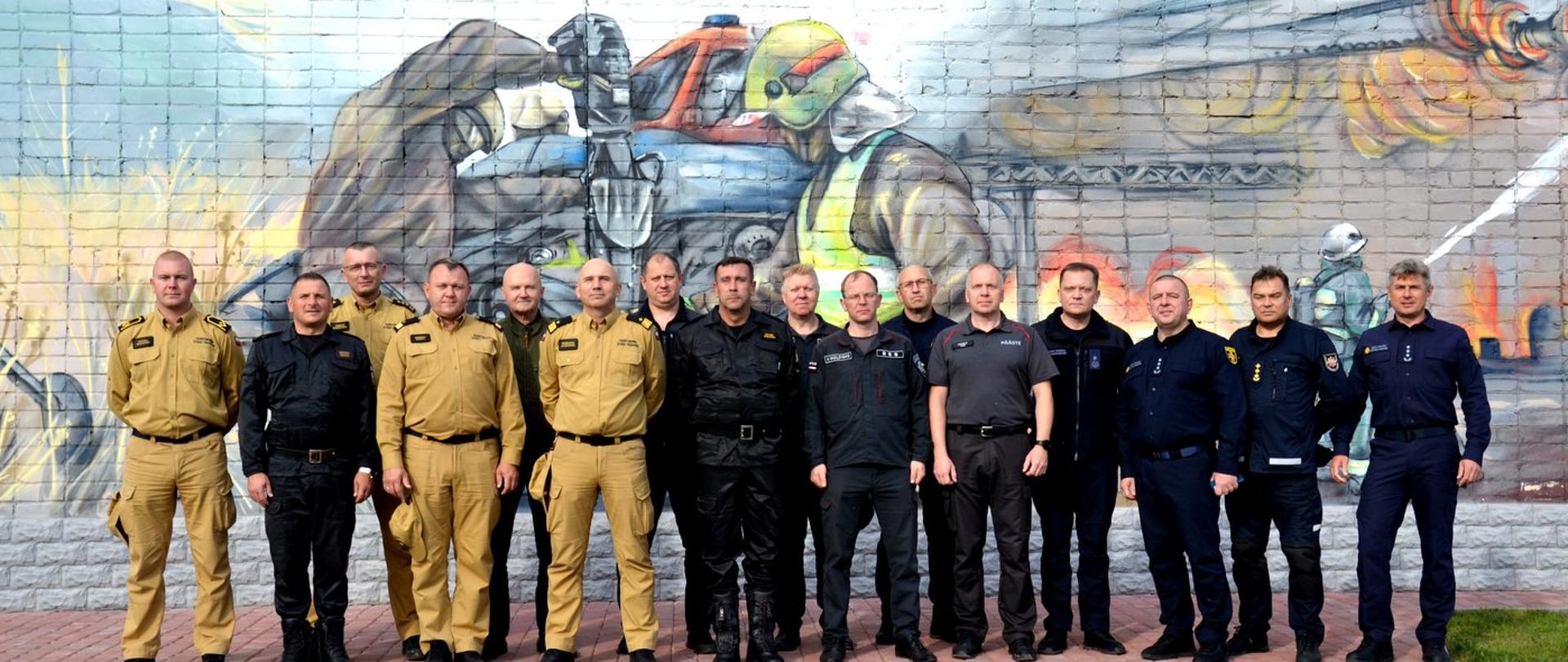 Zdjęcie zrobione na zewnątrz. Przed muralem przedstawiającym działania ratowniczo-gaśnicze w dwuszeregu stoi delegacja strażaków. Wśród nich strażacy Państwowej Straży Pożarnej oraz ratownicy z Ukrainy, Łotwy, Litwy i Estonii.
