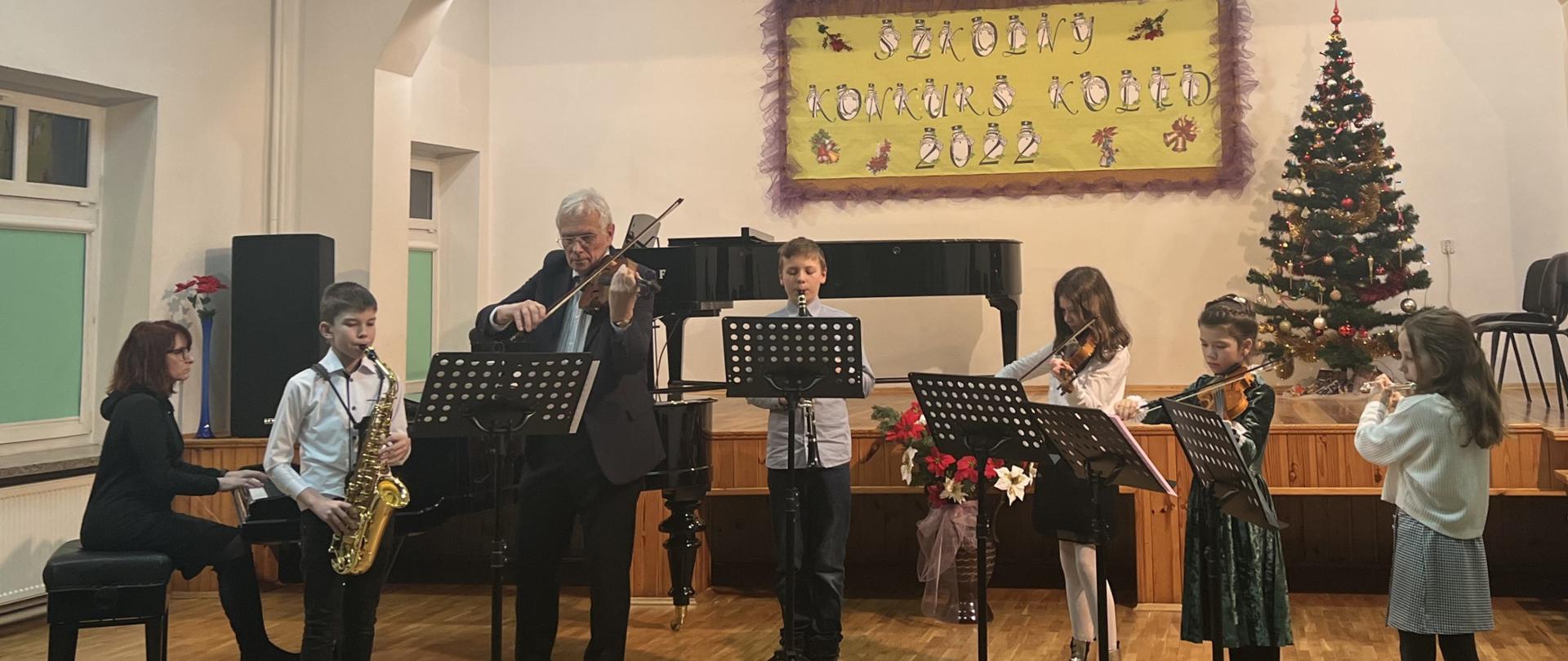 Zdjęcie przedstawiające zespół kameralny pod kierownictwem Tadeusza Wachowskiego, podczas szkolnego konkursu kolęd w auli szkoły z akompaniamentem fortepianowym Halyny Hachak.