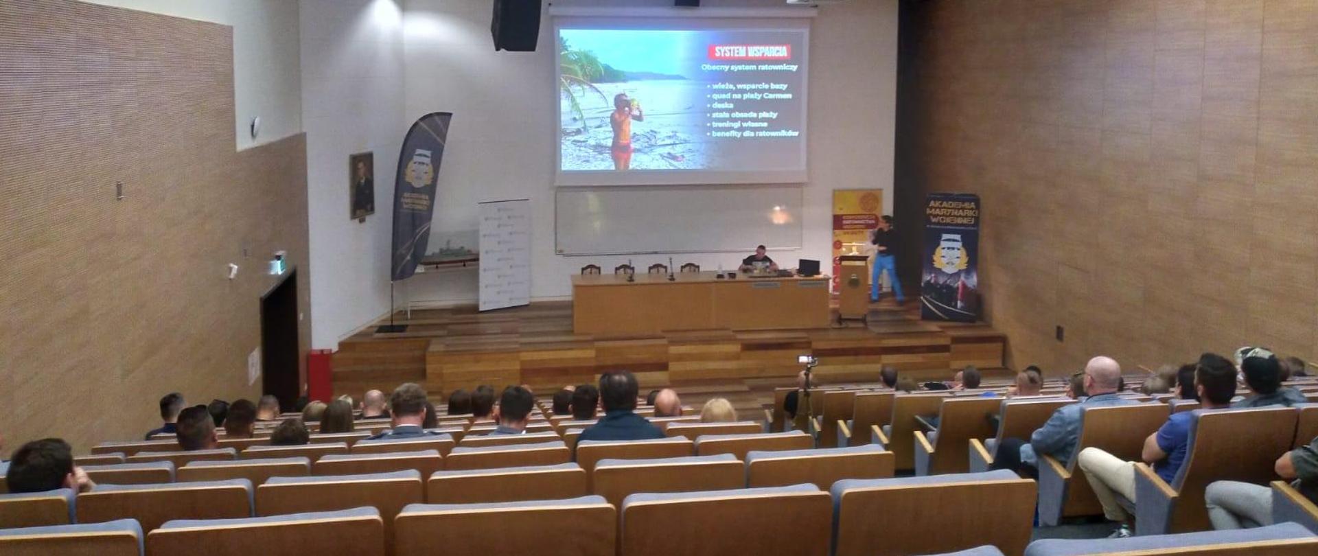Widok z tyłu na uczestników wykładu siedzących w auli, w głębi podest ze stołem konferencyjnym przy którym siedzi mężczyzna. Za nim na ścianie ekran multimedialny z wyświetlonym slajdem prezentacji