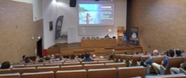 Widok z tyłu na uczestników wykładu siedzących w auli, w głębi podest ze stołem konferencyjnym przy którym siedzi mężczyzna. Za nim na ścianie ekran multimedialny z wyświetlonym slajdem prezentacji
