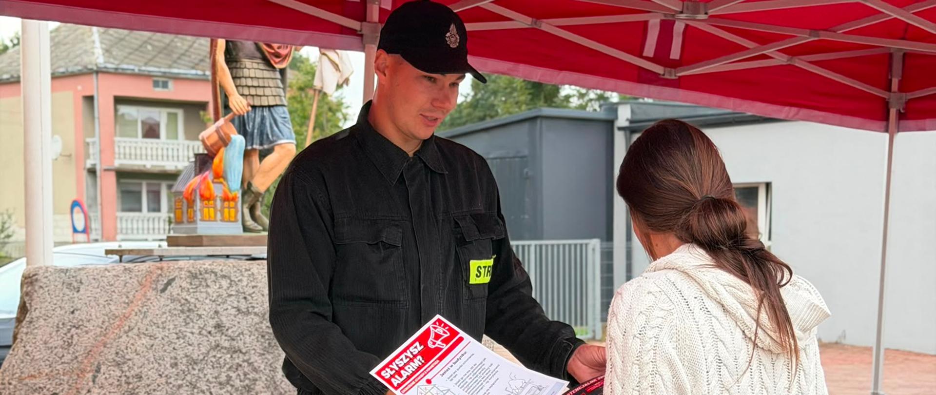 Strażak tłumaczący Pani Zasady Bezpiecznego Reagowania na wypadek zagrożenia. Oboje znajdują się pod czerwonym strażackim namiotem. W ręku kobieta trzyma ulotkę informacyjną. 
