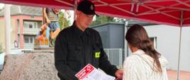 Strażak tłumaczący Pani Zasady Bezpiecznego Reagowania na wypadek zagrożenia. Oboje znajdują się pod czerwonym strażackim namiotem. W ręku kobieta trzyma ulotkę informacyjną. 