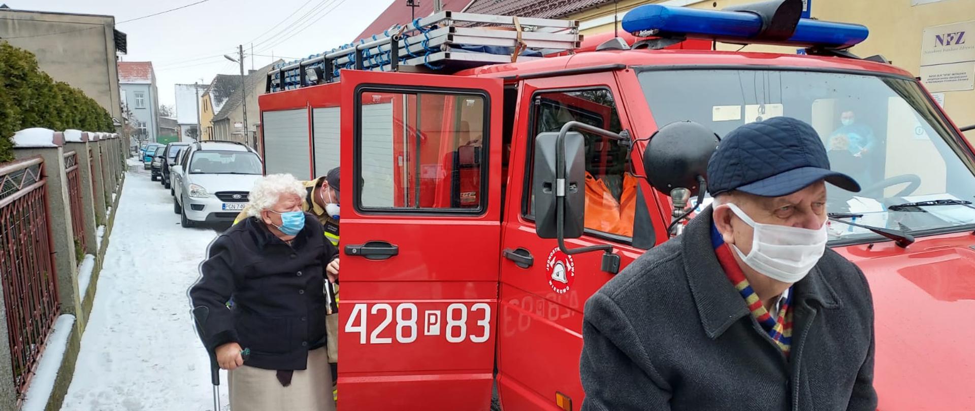 Na zdjęciu widać czerwony samochód strażacki. W samochodzie otwarte są tylne prawe drzwi. Przy drzwiach stoi starsza kobieta z laską w prawej ręce. Za nią widoczny jest lekko pochylony strażak, który pomagał seniorce wyjść z pojazdu. Na wysokości przedniego zderzaka idzie starszy pan w czapeczce z daszkiem. Wszyscy mają założone maseczki ochronne. Samochód stoi przy chodniku na którym widać leżący śnieg. W tle widać budynki oraz inne zaparkowane samochody osobowe.