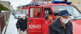 Na zdjęciu widać czerwony samochód strażacki. W samochodzie otwarte są tylne prawe drzwi. Przy drzwiach stoi starsza kobieta z laską w prawej ręce. Za nią widoczny jest lekko pochylony strażak, który pomagał seniorce wyjść z pojazdu. Na wysokości przedniego zderzaka idzie starszy pan w czapeczce z daszkiem. Wszyscy mają założone maseczki ochronne. Samochód stoi przy chodniku na którym widać leżący śnieg. W tle widać budynki oraz inne zaparkowane samochody osobowe