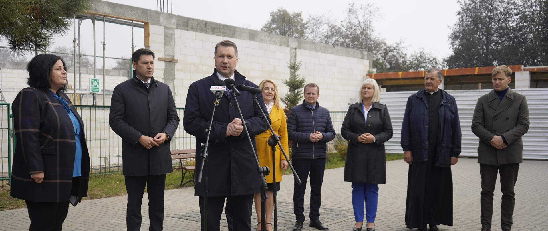 Na wyłożonym szarą kostką placyku stoi minister Czarnek i mówi do mikrofonu na stojaku, za nim stoi kilka osób, w tle rozpoczęta budowa.