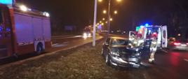 Pora nocna, na zdjęciu widać rozbity przód auta osobowego, który stoi na pasie zieleni. Samochód ma otwarte drzwi kierowcy a przy nich stoi trzech strażaków. Za samochodem po prawej stronie stoi karetka pogotowia, która ma otwarte tylne drzwi, widać w nich ratownika medycznego. Po lewej stronie zdjęcia widać samochód strażacki