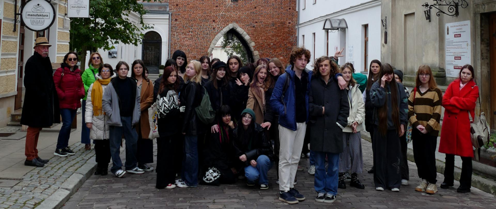fotografia barwna, na pierwszym planie grupa młodych osób, po lewej stronie mężczyzna w kapeluszu. na drugim planie znajduje się Brama Opatowska