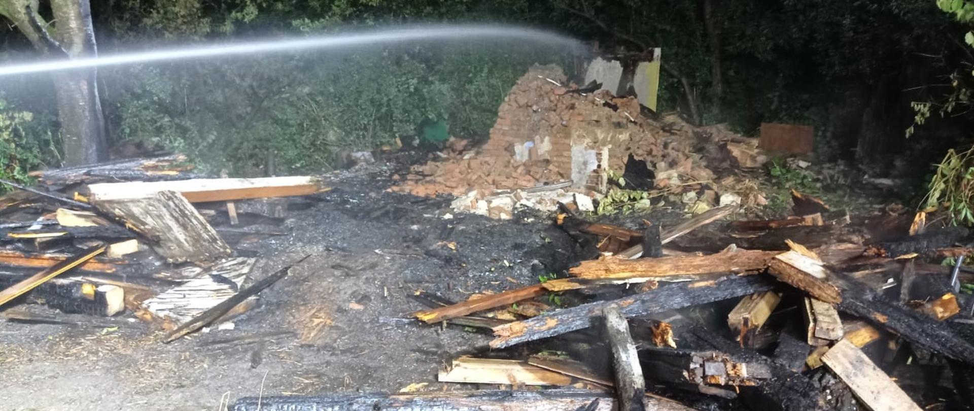Zdjęcie przedstawia zgliszcza po pożarze, prawdopodobnie budynku. Na pierwszym planie widoczne są zwęglone kawałki drewna i inne spalone resztki, leżące na ziemi. W tle widać stos gruzu i cegieł, będący prawdopodobnie pozostałością po ścianach budynku. Z lewej strony kadru, woda z węża strażackiego jest rozpylana, gasząc resztki pożaru. Otoczenie jest ciemne, z zarysem drzew i krzewów w tle, co sugeruje, że zdjęcie zostało wykonane w nocy lub o zmierzchu.