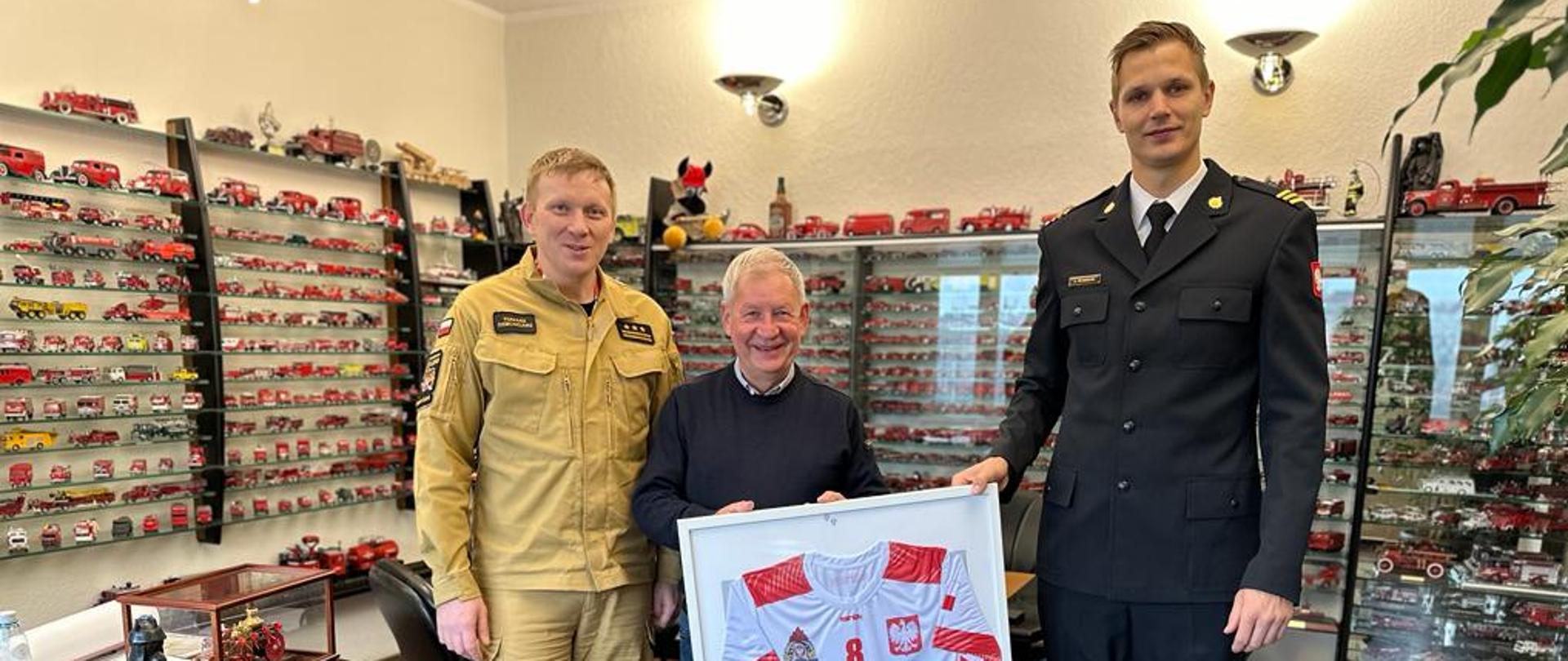 W gabinecie wiceprezesa Noma2 stoją komendant miejski st. bryg. Tomasz Dziechciarz, Wiceprezes Zarządu Spółki Noma2 Pan Marian Kurzal oraz sekc. Łukasz Żłobecki przekazujący pamiątkową koszulkę meczową z igrzysk.