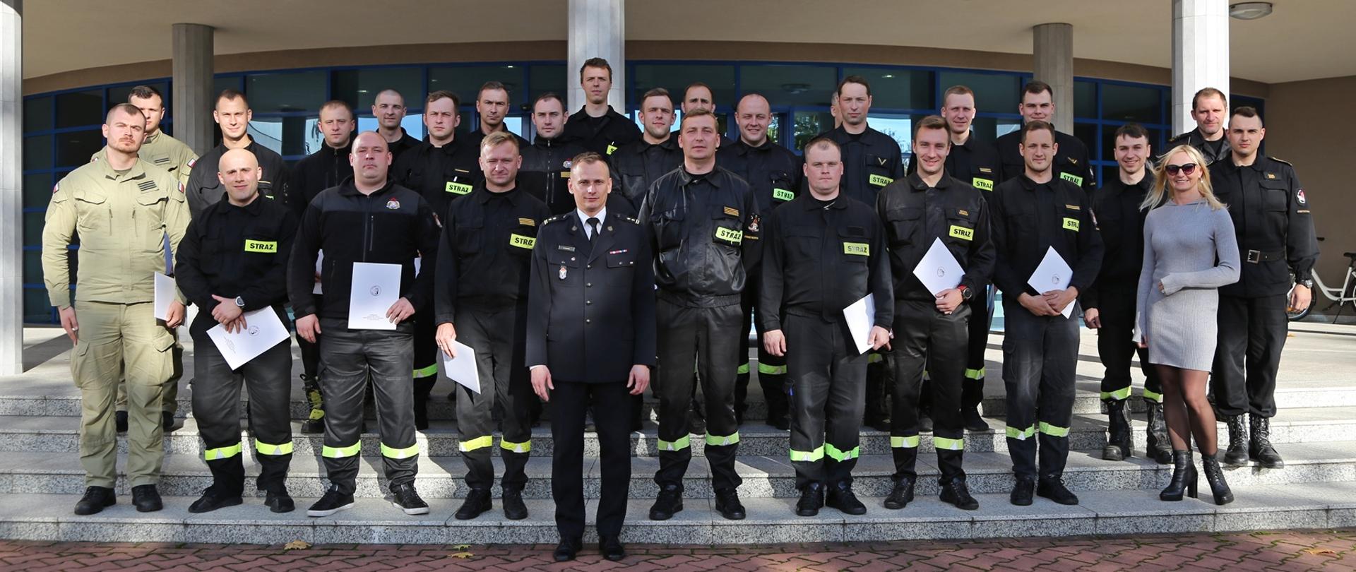
Zakończenie szkolenia ratownictwa chemicznego i ekologicznego - zdjęcie grupowe wszystkich uczestników szkolenia z dyplomami ukończenia kursu, przed nimi Zastępca Komendanta Szkoły