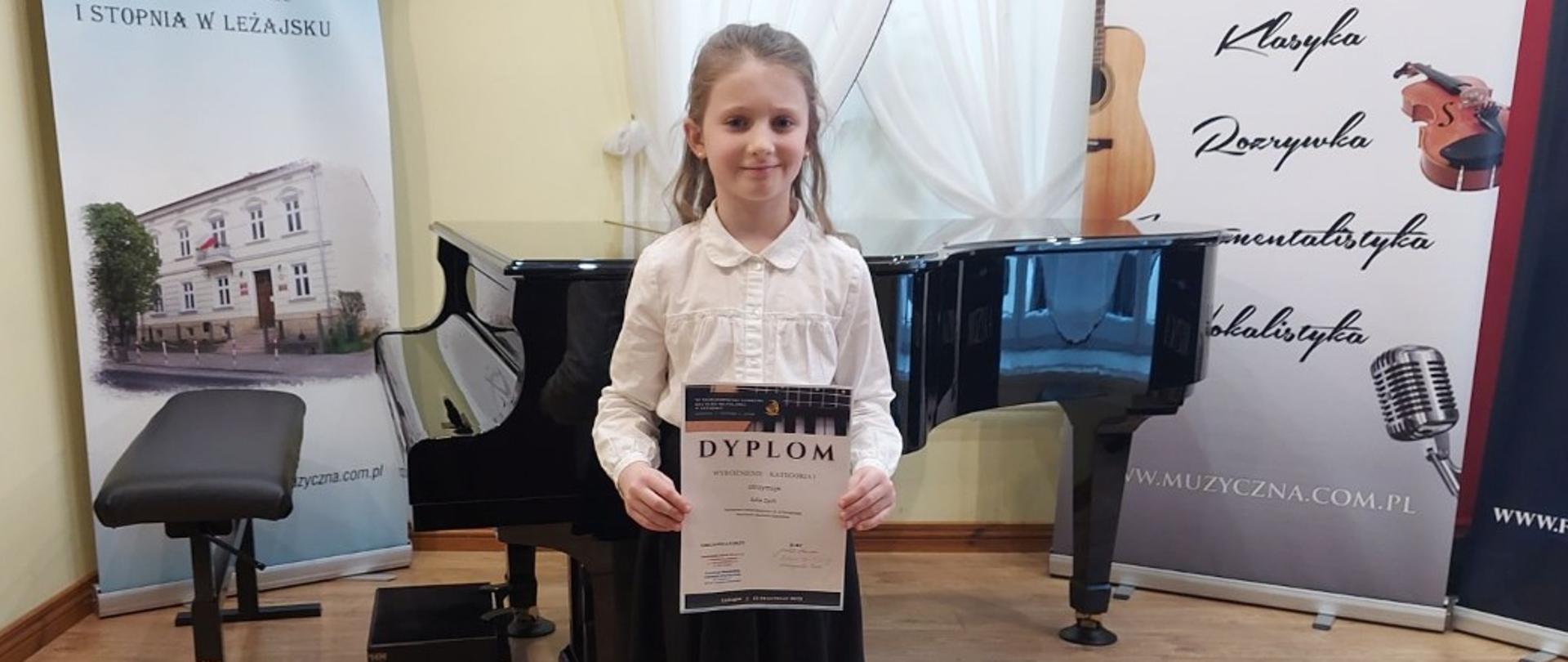 Zdjęcie przedstawia dziewczynkę trzymającą w rękach dyplom wyróżnienia. Z tyłu widoczny jest fortepian wraz z krzesłem oraz podnóżkiem.