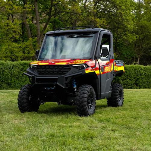 Terenowy pojazd typu UTV (quad z kabiną) marki Polaris Ranger 1000 w jaskrawych barwach strażackich – czerwonej z żółto-neonowymi pasami ostrzegawczymi i napisem „STRAŻ”. Pojazd posiada masywne opony z głębokim bieżnikiem i stoi na zielonej trawie.