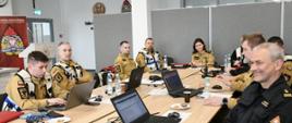 Funkcjonariusze Państwowej Straży Pożarnej siedzą przy stołach na których rozłożone są laptopy oraz radiostacje.