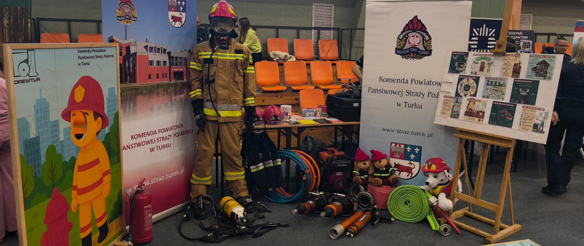 Stoisko edukacyjne Państwowej Straży Pożarnej w hali sportowej. Na środku stoi manekin ubrany w pełny strój strażaka z hełmem i maską. Wokół rozłożony jest sprzęt ratowniczy, m.in. węże, prądownice, butle powietrzne i gaśnica. Po lewej stronie widoczna jest plansza z ilustracją strażaka, a po prawej tablica edukacyjna. Na podłodze leży fantom do nauki pierwszej pomocy. W tle znajdują się trybuny z pomarańczowymi siedzeniami oraz uczestnicy wydarzenia.