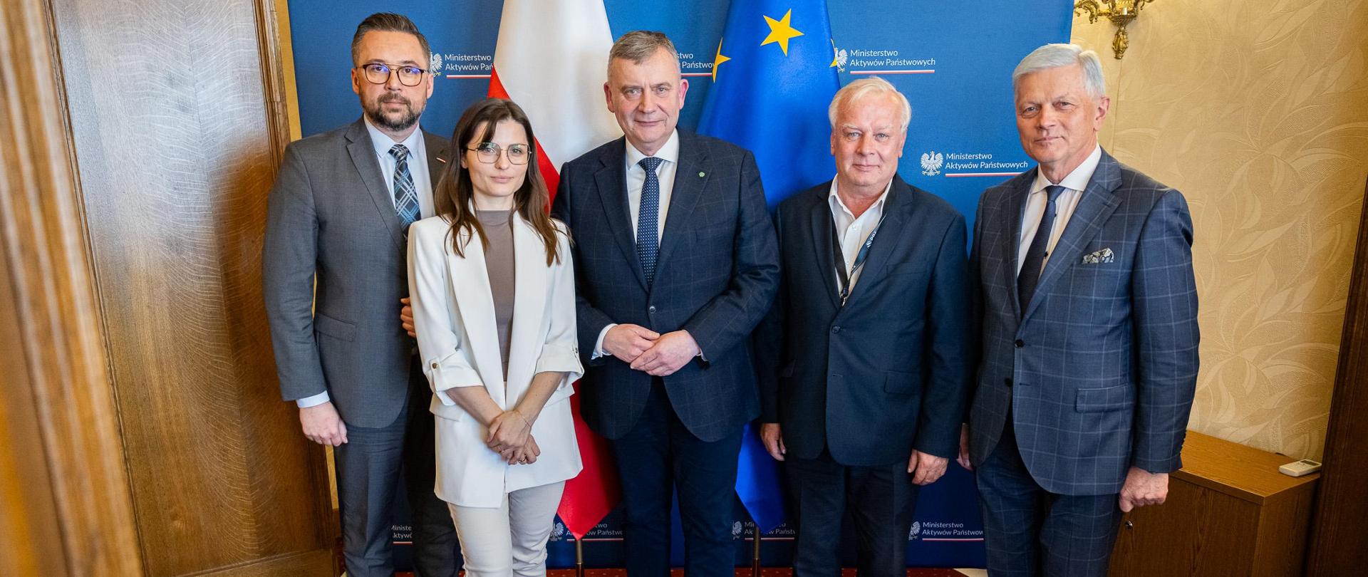 Na tle ścianki MAP oraz flagi Polski i Unii Europejskiej stoją od lewej Wiceminister MAP Marcin Kulasek, wojewoda Wielkopolska Agata Sobczyk, wiceminister MON Paweł Bejda, wiceminister MRiT Jacek Tomczak oraz Przewodniczący Komisji Obrony Narodowej Andrzej Grzyb. Po prawej stronie ścianki kinkiet, pod nim szafka.