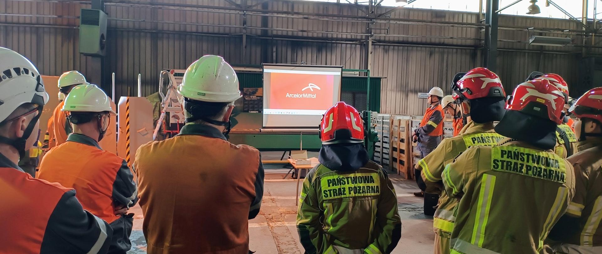 Zdjęcie przedstawia grupę strażaków oraz pracowników zgromadzonych w dużej hali przemysłowej na szkoleniu lub odprawie. Uczestnicy są odwróceni tyłem do obiektywu i patrzą w stronę wyświetlanej na ekranie prezentacji. Część osób ma na sobie piaskowe mundury strażackie z napisami na plecach, a pozostali noszą odzież roboczą z jaskrawymi, pomarańczowymi elementami odblaskowymi. Wszyscy obecni dbają o bezpieczeństwo, mając na głowach specjalistyczne kaski ochronne w kolorach czerwonym, białym i żółtym.
Scena rozgrywa się w przestronnym, surowym wnętrzu o wysokim suficie z widoczną konstrukcją stalową i systemem oświetlenia górnego. W tle widać okna wpuszczające naturalne światło oraz elementy wyposażenia warsztatowego, takie jak palety i skrzynie. Centralnym punktem uwagi jest stanowisko z monitorem lub rzutnikiem, przy którym prowadzący spotkanie przekazuje informacje techniczne. Całość kompozycji, z bliskim planem na ramiona i kaski osób stojących z przodu, nadaje zdjęciu perspektywę uczestnika wydarzenia.
