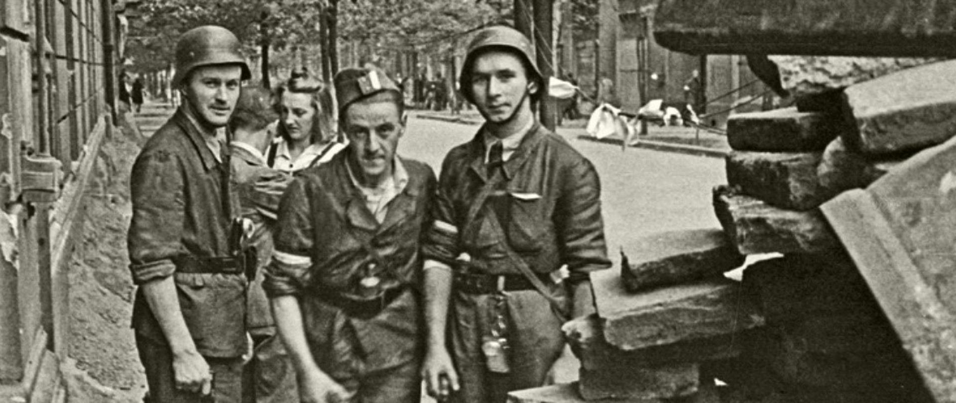 Trzech powstańców warszawskich stoi na tle ulicy.