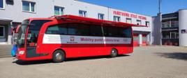 Zdjęcie przedstawia krwiobusa koloru czerwonego, który zaparkowany jest na placu przed budynkiem komendy