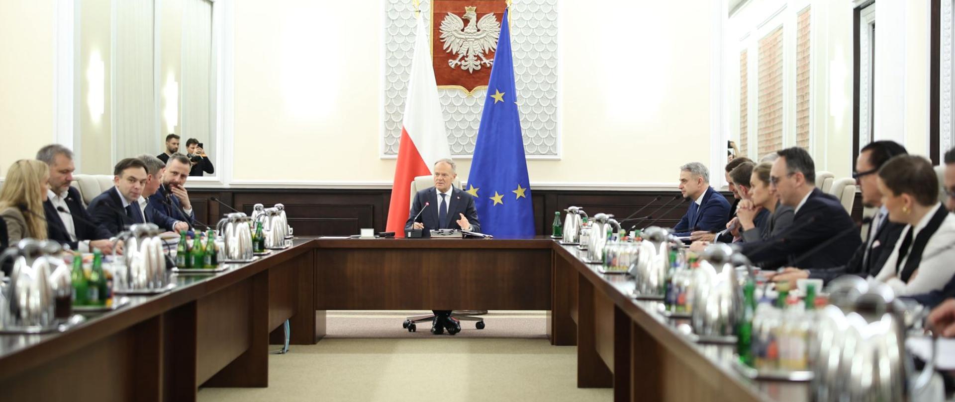 Zdjęcie od tyłu sali, za ustawionymi w czworokąt stołami siedzą ludzie, u szczytu stołu premier, za nim flagi Polski i UE, nad nimi na białej ścianie godło.