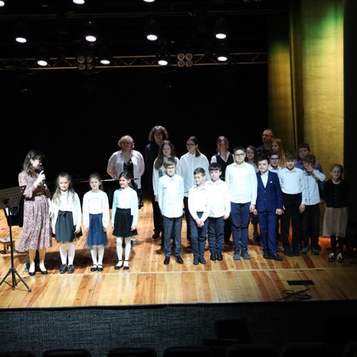 Fotografia wspólna wszystkich wykonawców koncertów dla uczniów szkół podstawowych. Ustawieni w rzędzie na scenie MOK w Głubczycach, przed oraz z prawej strony obok fortepianu.