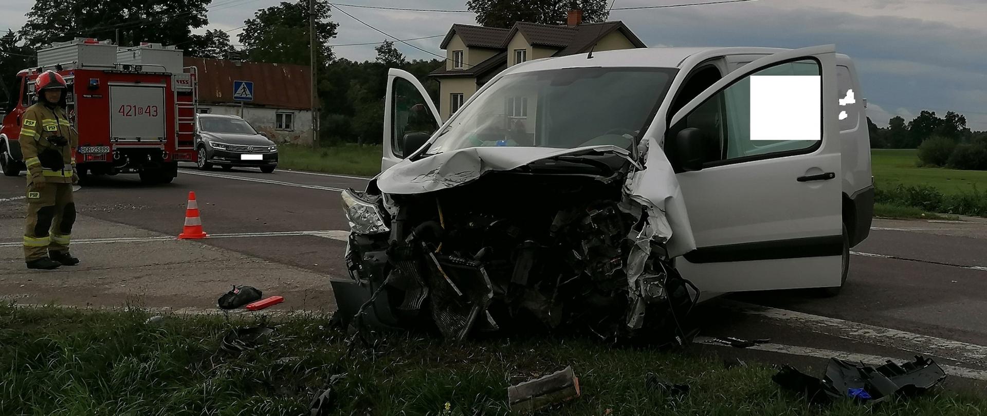 Rozbity samochód typu bus.Z lewej stoi strażak PSP za nim pojazd pożarniczy.