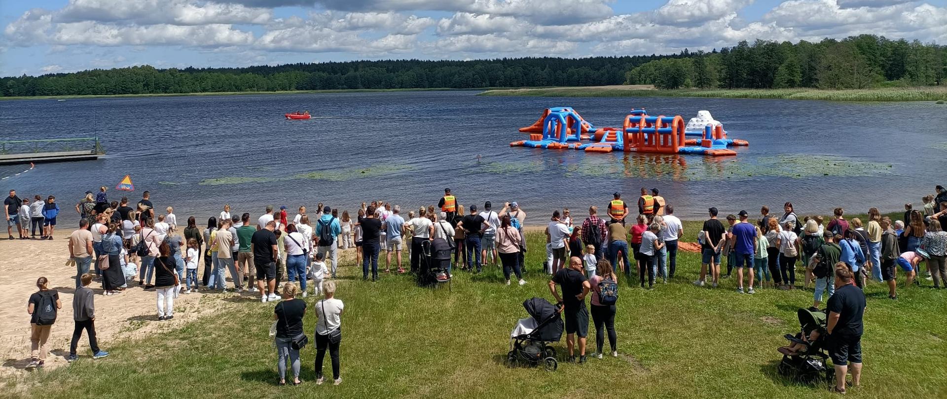 Zdjęcie przedstawia funkcjonariuszy Komendy Powiatowej Państwowej Straży Pożarnej w Gołdapi oraz dzieci, młodzież i osoby dorosłe uczestniczące w akcji "Bezpieczne Wakacje" na plaży miejskiej w Gołdapi. W tle widać strażaków płynących łodzią ratowniczą