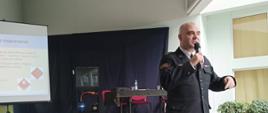 Strażak Państwowej Straży Pożarnej w mundurze wyjściowym przemawia przez mikrofon obok na stole stoi rzutnik multimedialny oraz ekran na którym wyświetlana jest prezentacja za nim ustawiony jest stół z krzesłami.