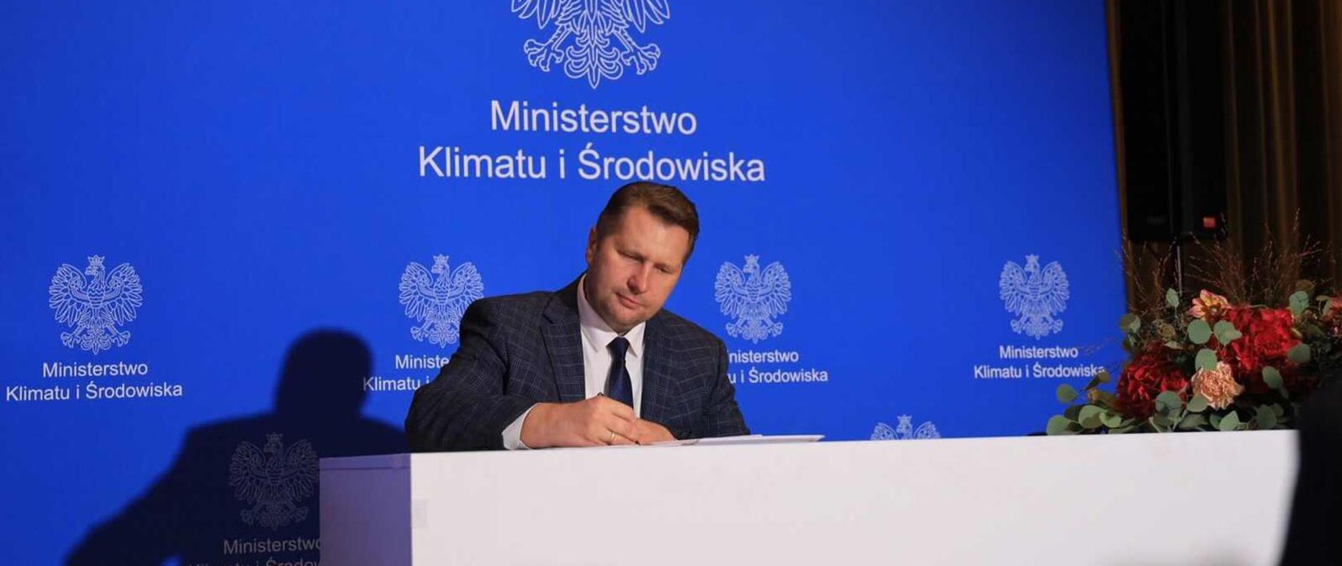 Minister Przemysław Czarnek podpisuje dokument siedząc przy białym stole. W tle niebieska ścianka z logo Ministerstwa Klimatu i Środowiska