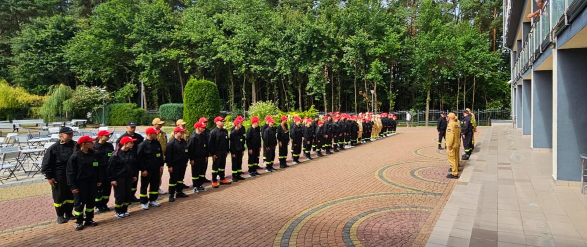 Na zdjęciu znajdują się dzieci stojące na baczność w czarnych strojach z czerwonymi czapkami a na przeciwko nich stoją opiekunowie i funkcjonariusze PSP. w tle widać zalesiony teren. 