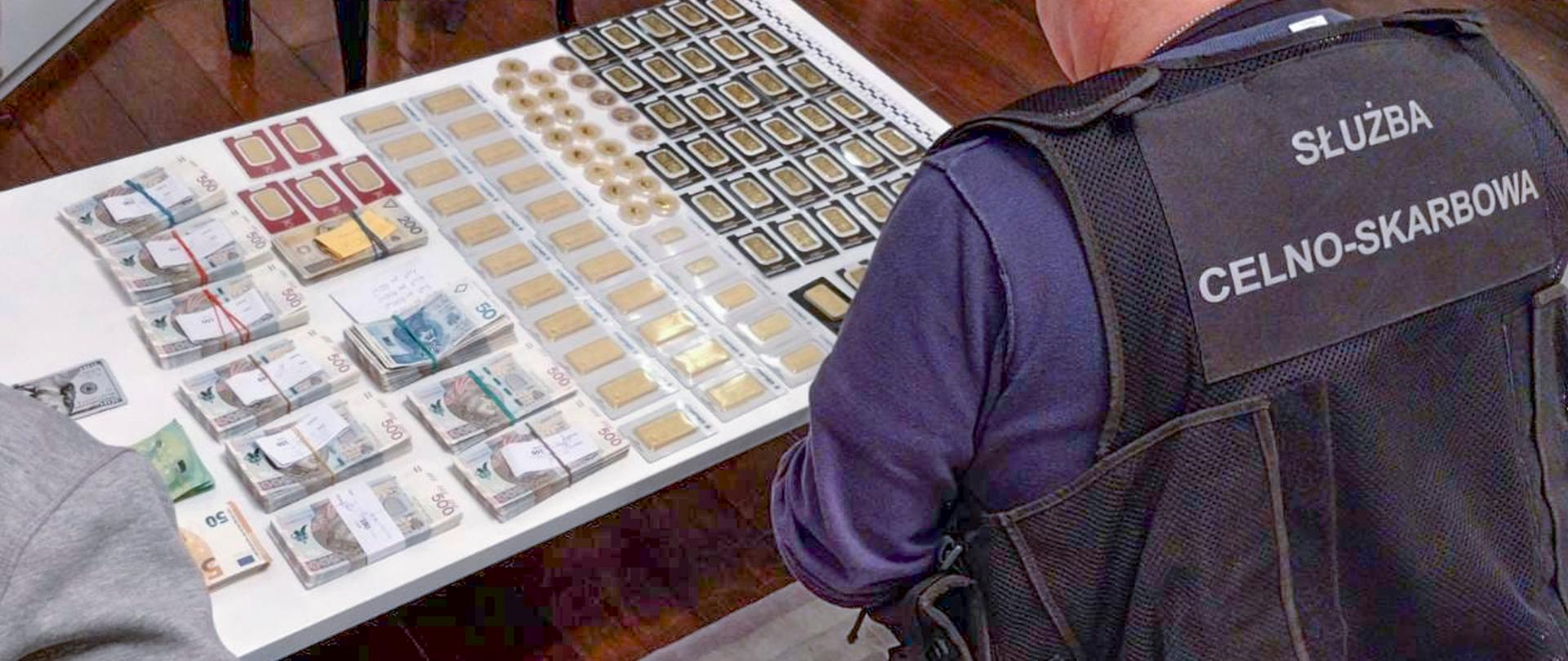 Dwóch funkcjonariuszy siedzi przy stole z plikami banknotów oraz dużą liczbą sztabek i monet.