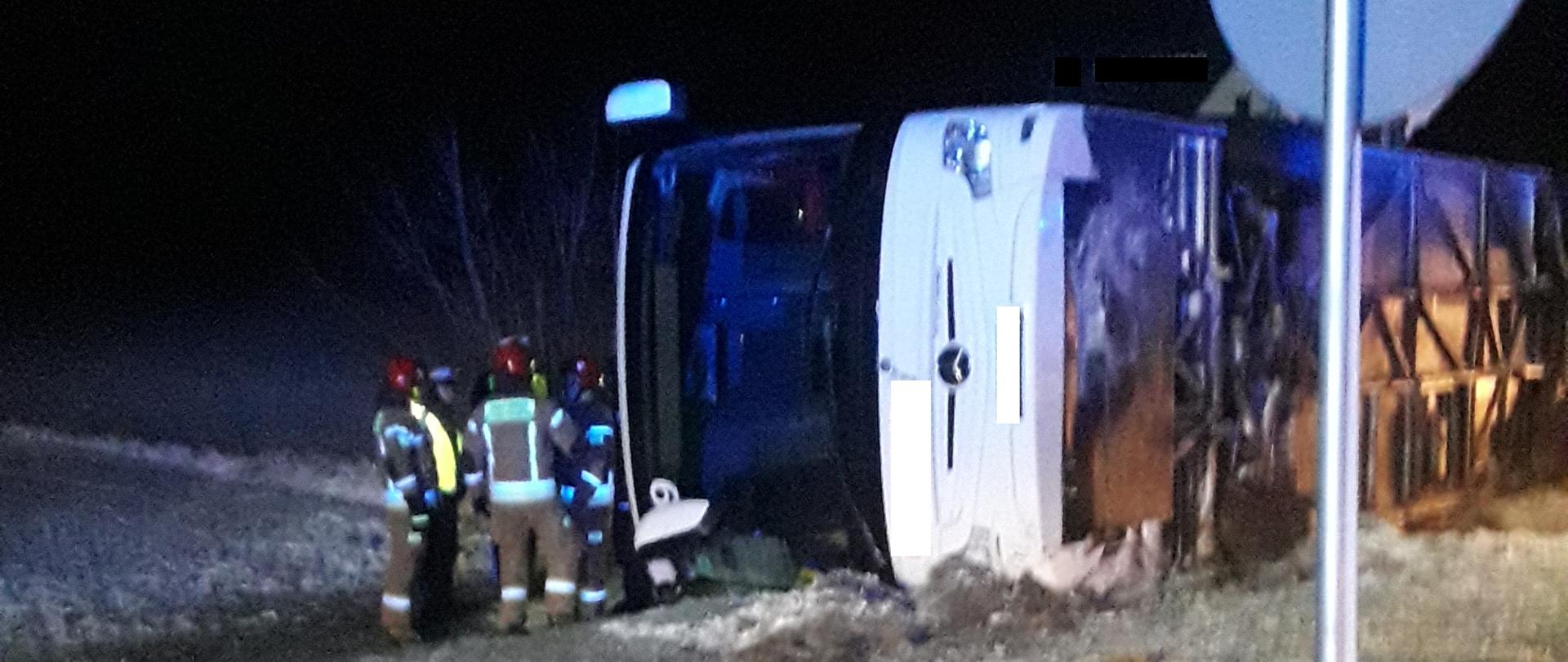 Autobus leży przewrócony na bok. Dookoła panuje zmrok. Przy autobusie pracują strażacy PSP i OSP. W tle pojazd straży pożarnej.