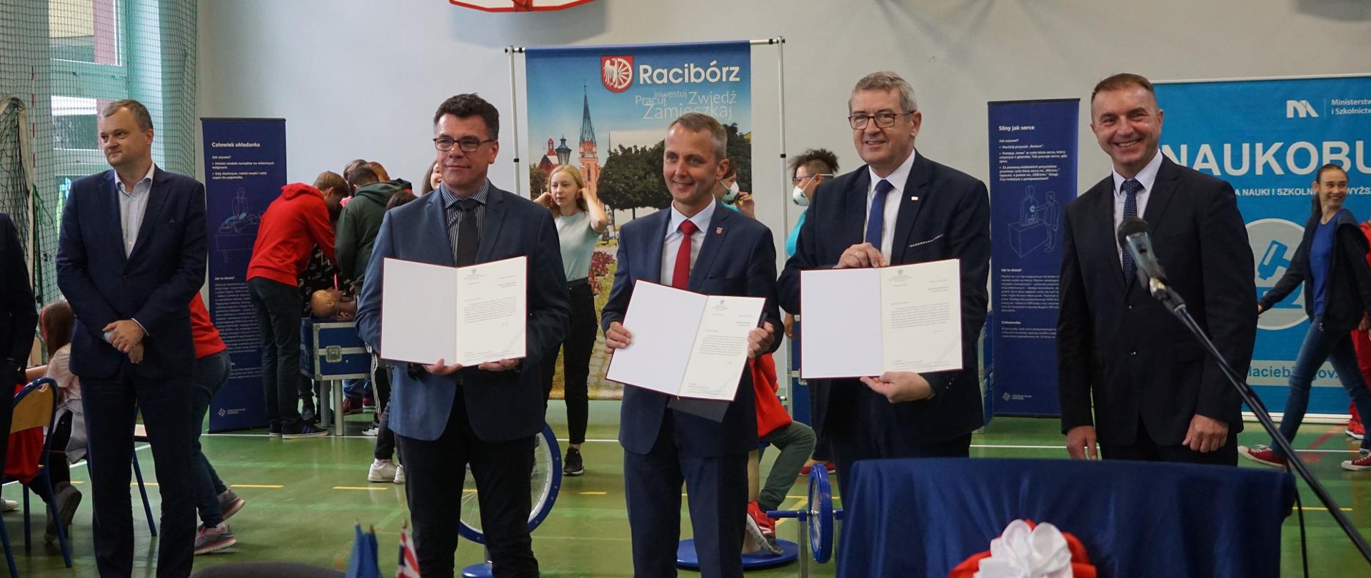 Minister i uczestnicy uroczystości prezentują podpisane dokumenty w sali gimnastycznej.