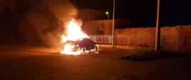 Na parkingu przy ogrodzeniu pali się samochód osobowy. Samochód pali się w całości, płomienie sięgają dwóch metrów. W bliskiej odległości znajduje się budynek.