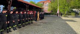 Strażacy w mundurach koloru czarnego oraz musztardowego stoją w rzędzie obok siebie przed wiatą strażacy w mundurze reprezentacyjnym stoją obok masztu i wciągają flagę Polski na maszt.
