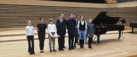 Ośmiu uczniów stoi przed dwójką nauczycieli przy fortepianie na scenie sali koncertowej PSM
