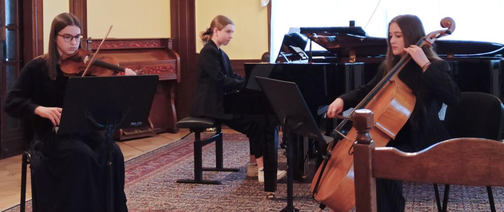 Zdjęcie kolorowe. Trzy uczennice w czarnych strojach - skrzypaczka, wiolonczelistka i pianistka - podczas występu.