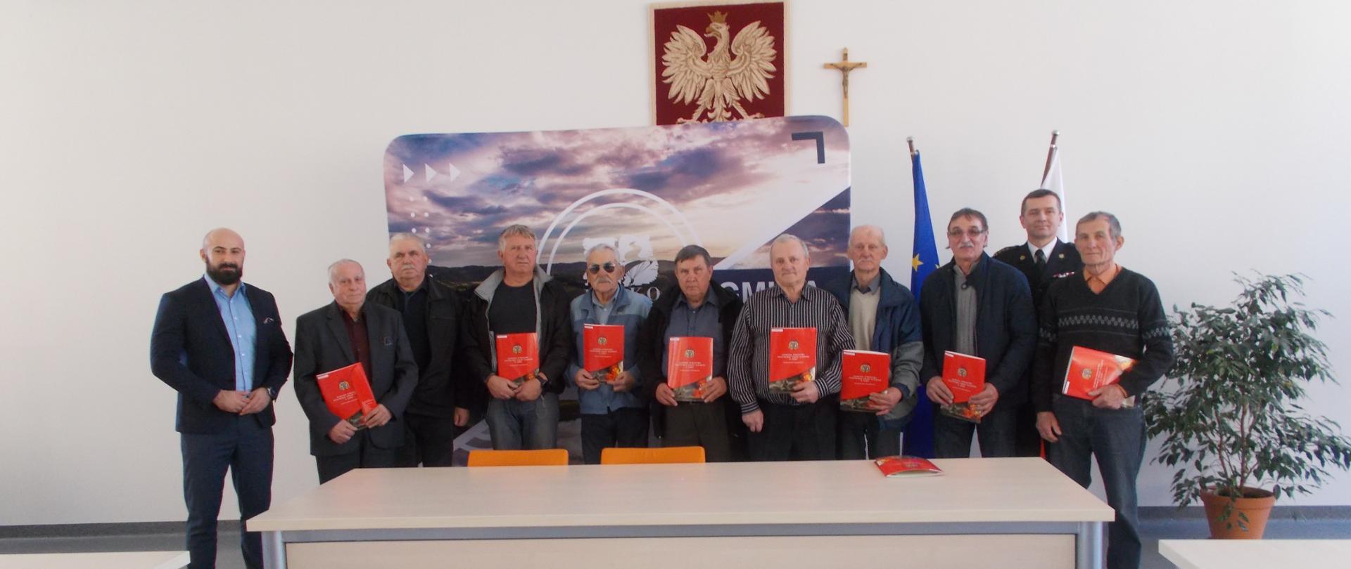 Zdjęcie przedstawia Burmistrza Leska Pana Adama Snarskiego oraz Komendanta Powiatowego Państwowej Straży Pożarnej w Lesku st. bryg. Jacka Dydka wraz z druhami Ochotniczych Straży Pożarnych z terenu Gminy Lesko. Każdy z druhów trzyma w rękach decyzję o przyznaniu świadczenia ratowniczego. 