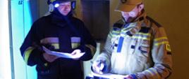 Widok z przodu. Po prawej stronie egzaminator – oficer PSP wpisuje do arkusza ocenę z zadania praktycznego. Po lewej stronie w ubraniu specjalnym i hełmie stoi druh OSP.