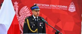 Za mównicą stoi komendant podczas przemówienia, ubrany jest w mundur galowy na głowie ma czapkę rogatywkę. Z tyłu biało-czerwona flaga oraz czerwony baner Państwowej Straży Pożarnej 