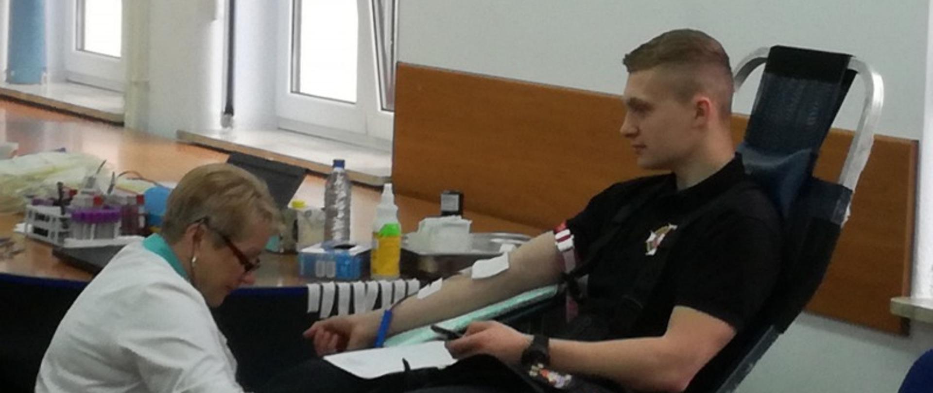 Na zdjęciu widoczny strażak w czarnej koszulce siedzący na fotelu i oddający krew po jego lewej stronie w białym fartuchu pielęgniarka.