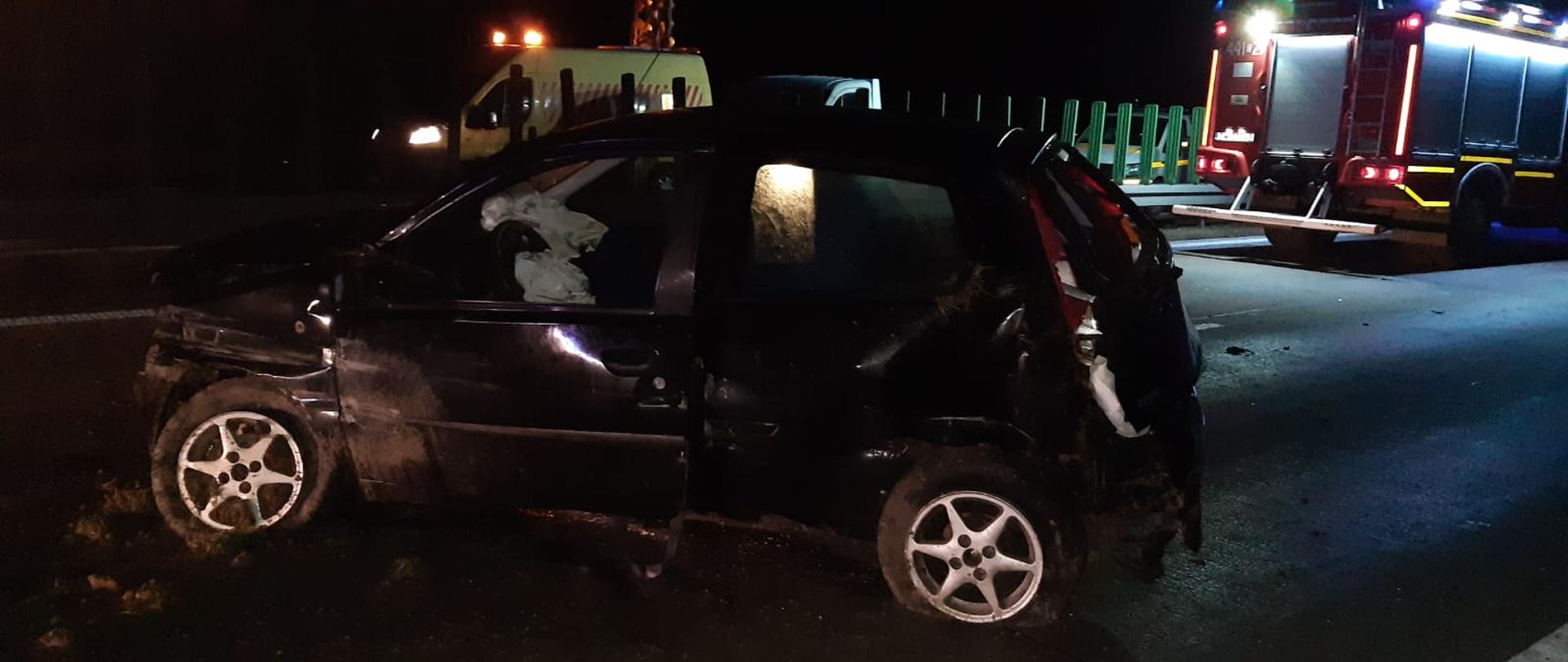 Obraz przedstawia pojazd osobowy po wypadku. W tle samochód pożarniczy oraz samochód zarządcy drogi. Pora nocna.
