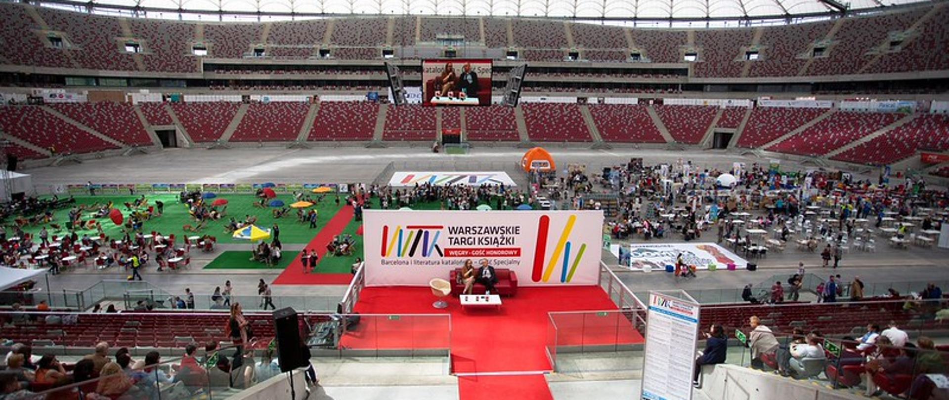 The arena of the PGE National Stadium; in the foreground, a large poster with the words "Warsaw Book Fair" written on it. The walkway to the poster is lined with red carpet; in front of the poster is a red couch on which a woman and a man are sitting and talking; behind the poster, the stadium arena is filled with people and stands.