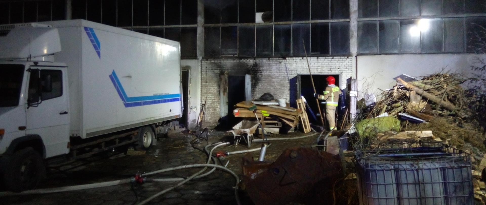  Na zdjęciu widoczny jest strażak trzymający w ręku bosak. W tle widać pozostałości wyposażenia magazynu, które zostały wyniesione na zewnątrz.