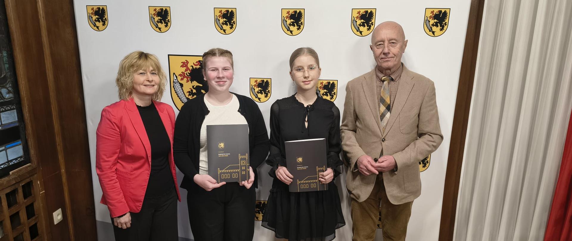 Cztery osoby stoją obok siebie w sali urzędowej. Dwie młode osoby na środku trzymają eleganckie teczki z herbem miasta. Po ich bokach stoją dorośli: kobieta w czerwonej marynarce oraz mężczyzna w jasnej marynarce i krawacie. W tle znajduje się ścianka z powtarzającym się herbem miasta, a podłoga jest drewniana.