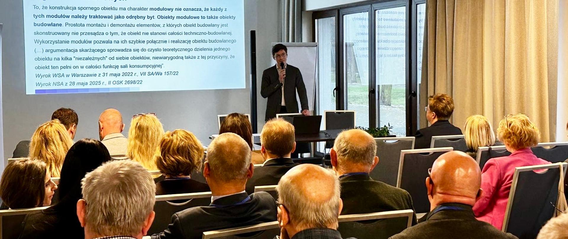 Filip Prusik-Serbinowski, Zastępca GINB podczas wystąpienia na naradzie szkoleniowej organów nadzoru budowlanego województwa łódzkiego. Na sali uczestnicy, w tle ekran z prezentacją