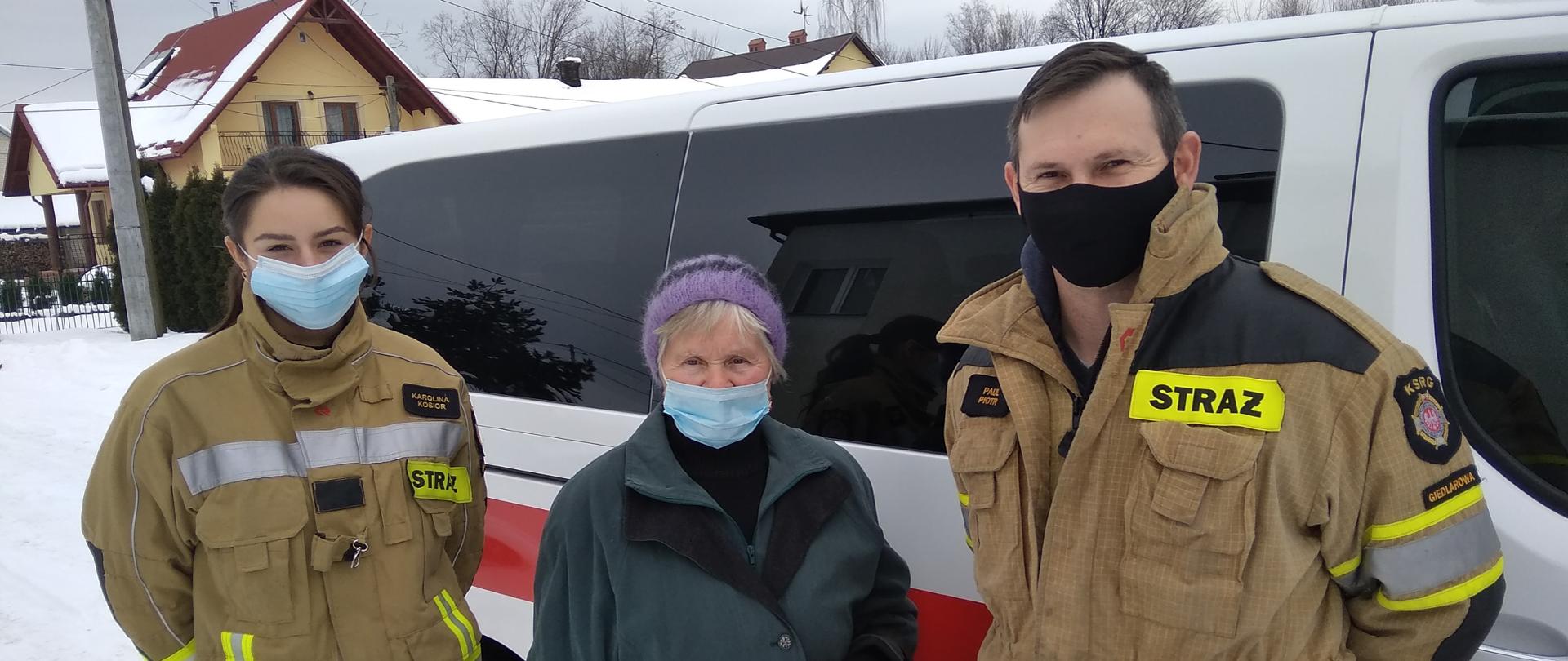 Na zdjęciu widzimy druhów ochotniczej straży pożarnej stojących po lewej i prawej stronie osoby starszej. W tle widzimy samochód oznakowany straży typu bus. 